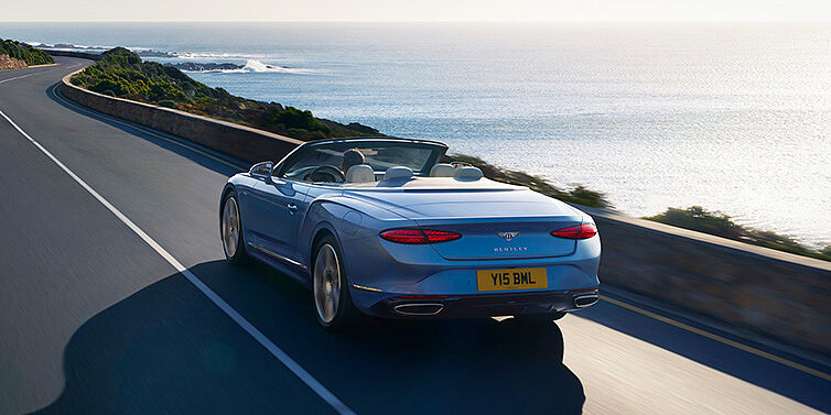 Gohm Sportwagen GmbH | Bentley Stuttgart New Bentley Continental GTC Azure convertible rear view in Light Windsor blue paint with 22 inch ten spoke wheels driving dynamically on an ocean road in the sunshine